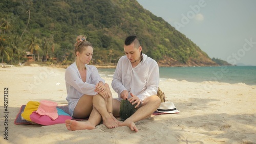 The cheerful love couple holding and eating slices of watermelon on tropical sand beach sea. Romantic lovers two people caucasian spend summer weekend in outdoor. Hat, backpack white shirt beachwear. © ivandanru