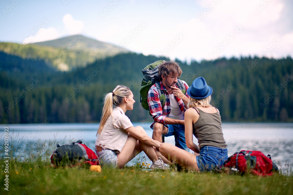 Friends in front of lake looking at map. Backpacker showing map to girls sitting in grass. Hiking in nature. Lifestyle, togetherness, nature concept