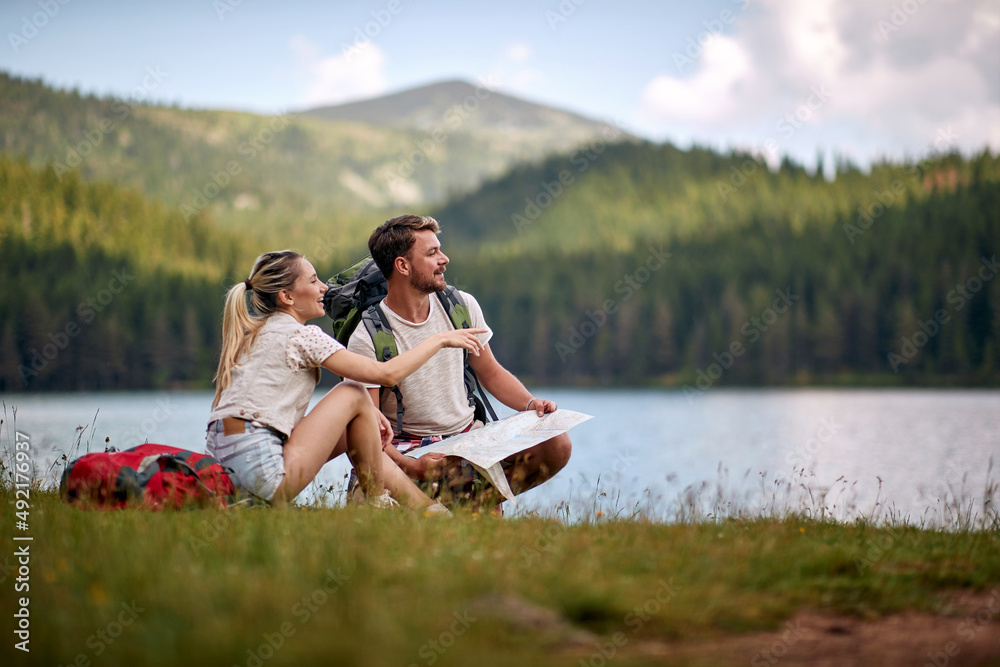 Tourist couple with map looking at lake. Blonde woman pointing to distance. Summer trip in nature. Lifestyle, togetherness, nature concept Tourist couple with map looking at lake. Blonde woman pointing to distance. Summer trip in nature. Lifestyle, togetherness, nature concept