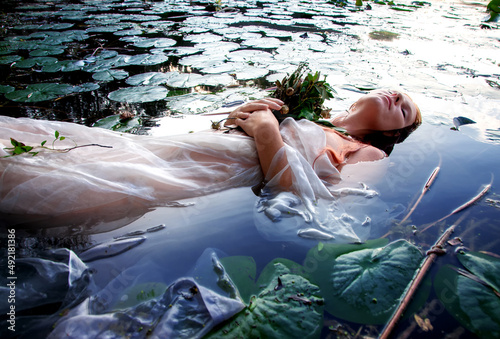 Young sexy woman Lady Ophelia lies with rotten flowers, eyes closed in white dress in the water between water lilies, in the lake photo