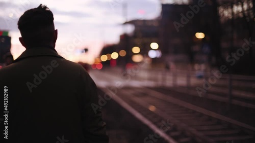 Back view of unrecognizable male silhouette in a coat moving along the tram rails the city center street at night.