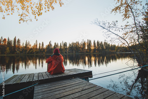 Pensive girl in a red jacket sits at the end of a wooden pier looking out into the calm water as the sun sets in Sotkamo, kainuu region, Finland. Scandinavia landscape photo