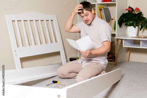 A puzzled, tired father cobbles the baby bed, wipes the sweat off his face. Heavy assembly of home furniture by yourself
