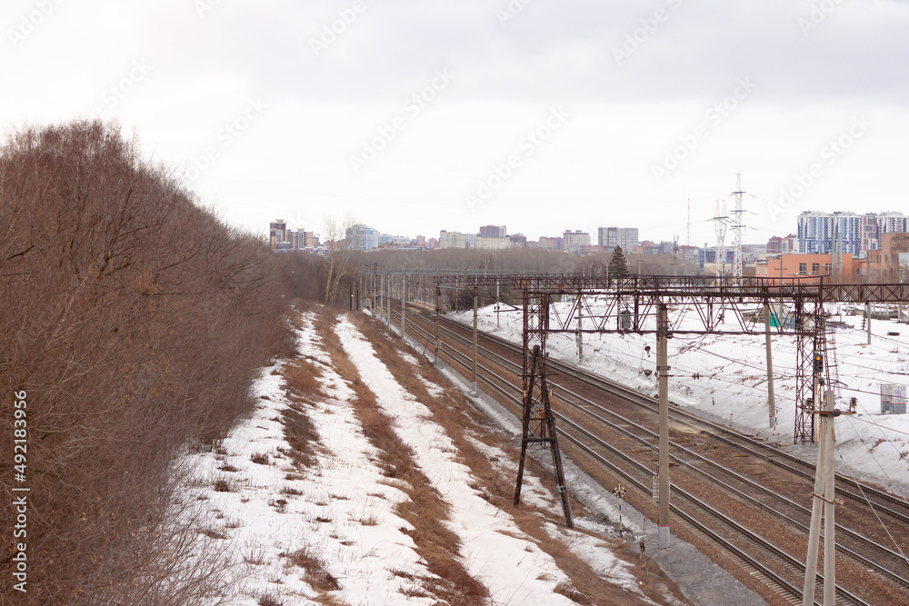 Railways leading up to the perspective to the city in the far away background. Leafless trees are on the side of the tracks and steel frame is on top of the rails