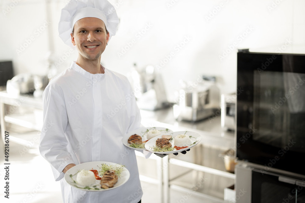 Portrait of young handsome smiling chef in uniform standing with ready meals in the kitchen. Happy caucasian cook working at restaurant. Concept of professional occupation and haute cuisine Portrait of young handsome smiling chef in uniform standing with ready meals in the kitchen. Happy caucasian cook working at restaurant. Concept of professional occupation and haute cuisine