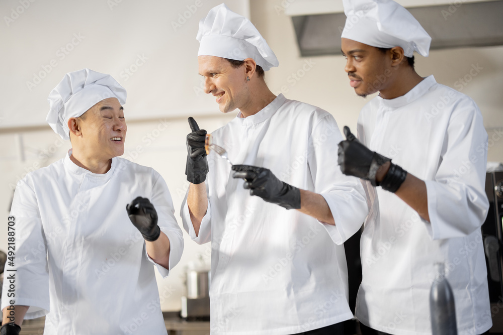 Three cooks with different nationalties tasting cooked meals, inventing new dishes for a menu. Caucasian chef with Latin and Asian cooks in uniform working together. Concept of teamwork and high