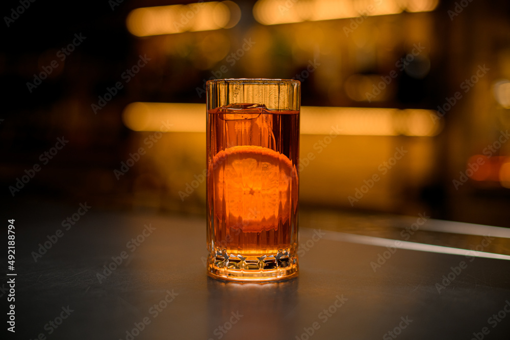 front view of tall glass full of cold alcoholic drink with orange slice on bar counter