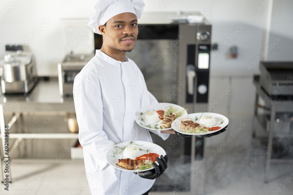 Portrait of young handsome smiling chef in uniform standing with ready meal in the kitchen. Happy Latin-American cook working at restaurant. Concept of professional occupation and haute cuisine Portrait of young handsome smiling chef in uniform standing with ready meal in the kitchen. Happy Latin-American cook working at restaurant. Concept of professional occupation and haute cuisine