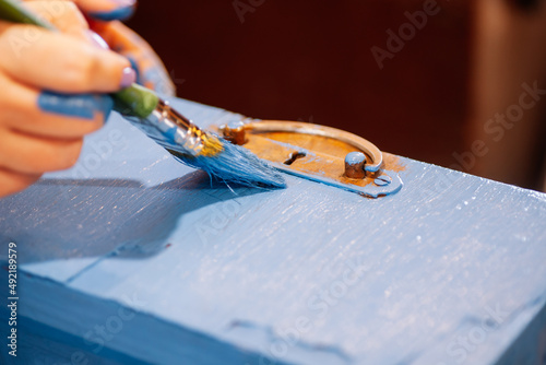 Closeup of paint-stained hands with big brush painting aged wooden closet drawer in blue color. Reuse of old things. Sustainable eco-friendly actions for planet future. 