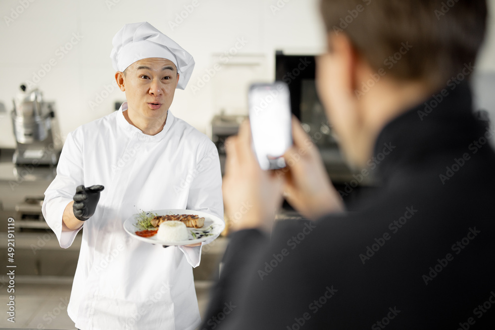 Asian chef in uniform talks about the cooked dish while maintains a culinary blog in professional kitchen. Man record him on phone. Concept of culinary video blogging Asian chef in uniform talks about the cooked dish while maintains a culinary blog in professional kitchen. Man record him on phone. Concept of culinary video blogging