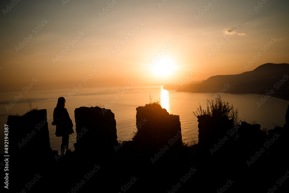 A woman or a man standing on a rock and looking directly at the sunset by the sea. The concept of nature and beauty. Orange sunset. Silhouette at sunset