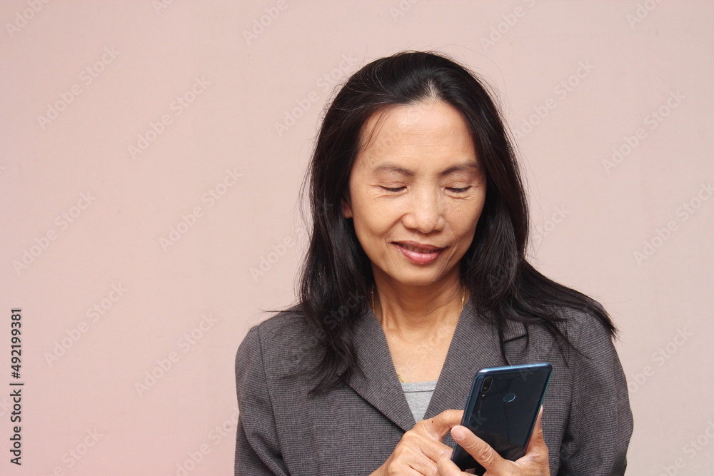 Portrait of an Asian business model holding a cell phone and looking at it with a relaxed mood. Wearing a gray suit against a light brown background.