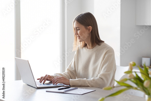 A young beautiful woman working on a laptop, sitting in the kitchen at home, remote work, freelance.
