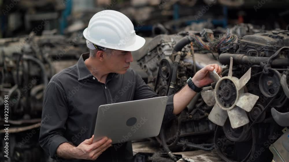 senior engineer using laptop computer checking list old used car engine in machinery parts shop warehouse. old technician man Repair vehicle automobile machine in Recycle motor factory