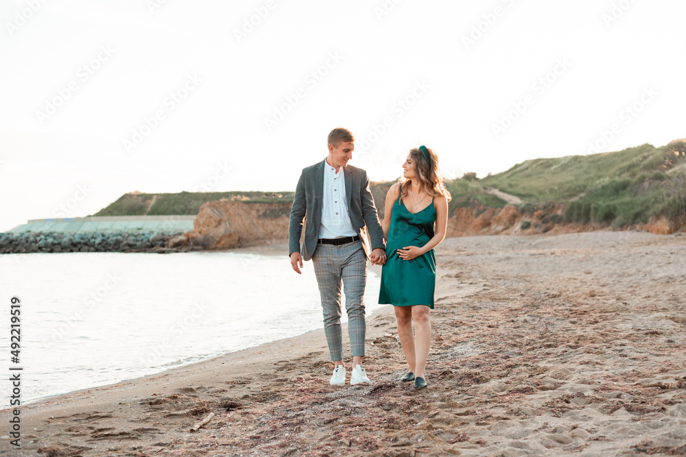Beautiful brunette pregnant woman with her husband walking hand by hand background sea Beautiful brunette pregnant woman with her husband walking hand by hand background sea