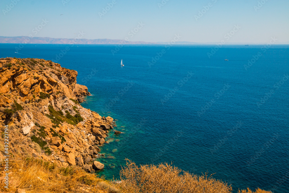 Fototapeta premium View from Cape Sounion (Greece) on Mediterranean Sea looking east with a sail boat in the background and the orange sand rocks in the foreground