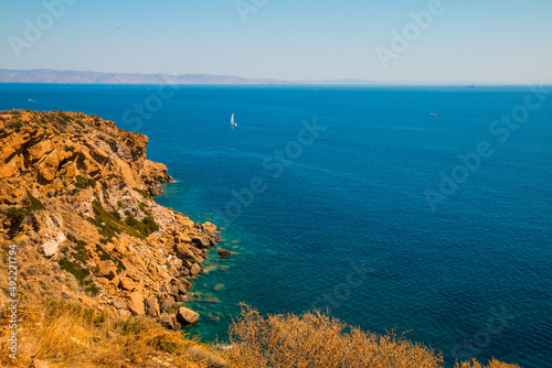 View from Cape Sounion (Greece) on Mediterranean Sea looking east with a sail boat in the background and the orange sand rocks in the foreground