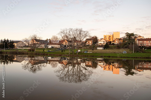 Sunset on a lake in Burgundy, France, with trees in winter and houses and buildings behind © sissoupitch