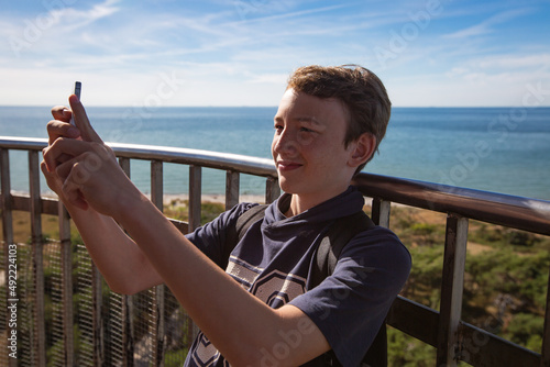 Young caucasian teenager on lighthouse takes selfie with cell phone - side view