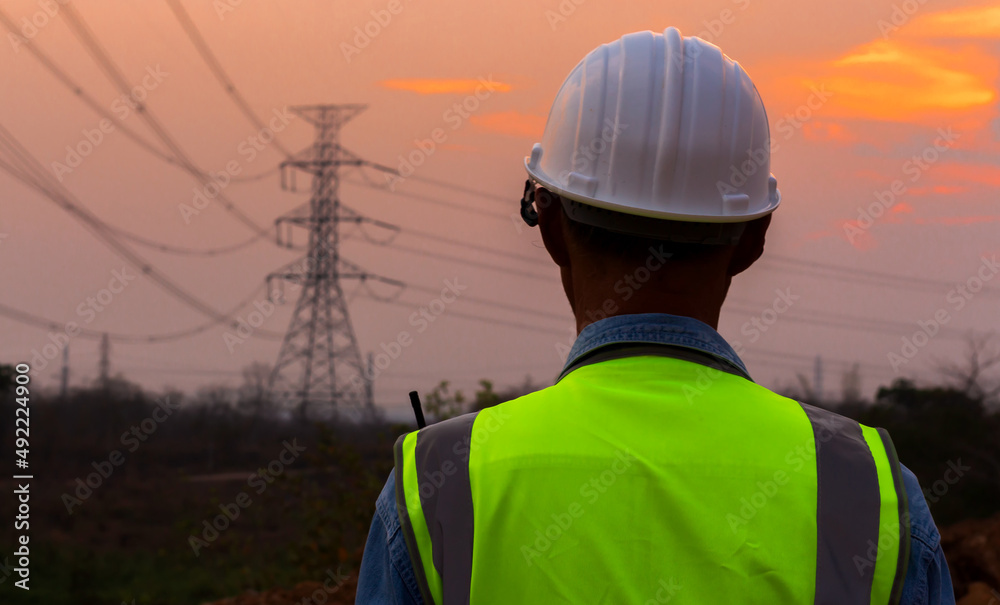 Rear view of an engineer wearing a hard hat in front of a high-voltage electric pole at dusk. Rear view of an engineer wearing a hard hat in front of a high-voltage electric pole at dusk.