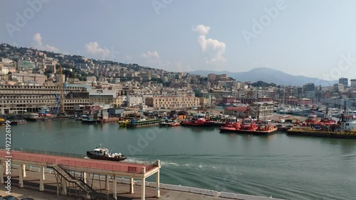 La Spezia port, Italy. A row of anchored boats, many of them tugboats. A small boat passes by. Horizon of hills with houses and green areas. Clear skies with a couple of vertical cloud formations. photo