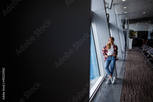 A beautiful attractive ukrainian woman with blond hair is standing by a large window, wearing momom jeans and a t-shirt with a plaid shirt. Girl in casual comfortable clothes