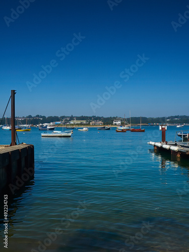 A view from a slipway across Bembridge Harbour, with yachts and boats floating on the mirror-like waters ahead of wooded hills and under blue skies.