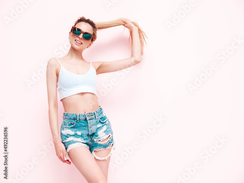 Young beautiful smiling female in trendy summer jeans shorts. Sexy carefree blond  woman posing near pink wall in studio. Positive model having fun. Cheerful and happy in sunglasses. Isolated © halayalex