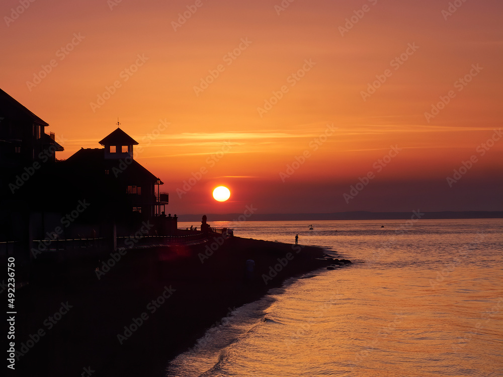 A richly coloured sunset seen from Cowes Beach, sunlight coming through the windows of a seaside building and  unrecognisable silhouetted people on the beach.