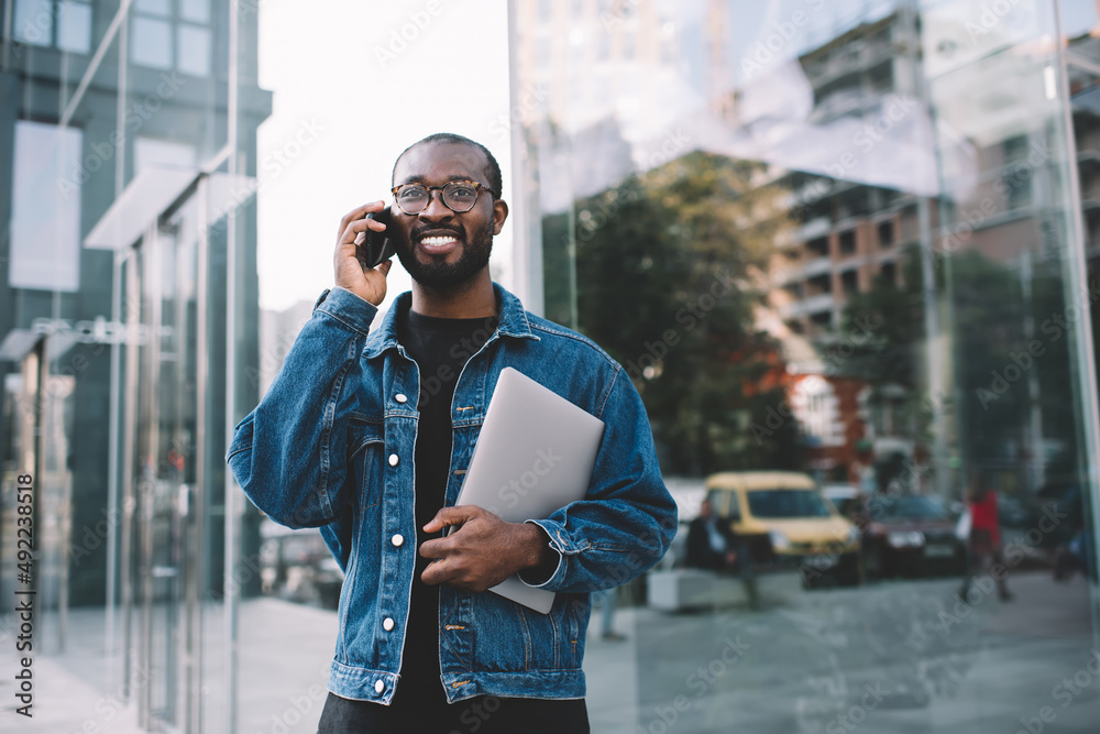 Cheerful African American male in classic spectacles for vision correction enjoying positive cellular conversation,happy dark skinned hipster guy with netbook making international call communication