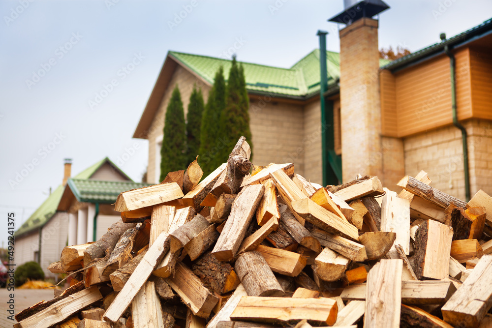 A pile of split firewood for heating the house, unloaded in the yard, against the backdrop of the house, natural heating sources. A pile of split firewood for heating the house, unloaded in the yard, against the backdrop of the house, natural heating sources.