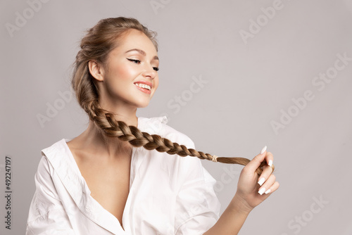 Happy pretty young woman holding her long pigtail, pleased with healthy shining hairs posing on gray background. Beauty portrait of smiling girl with stylish braid. Haircare treatment products ad. photo