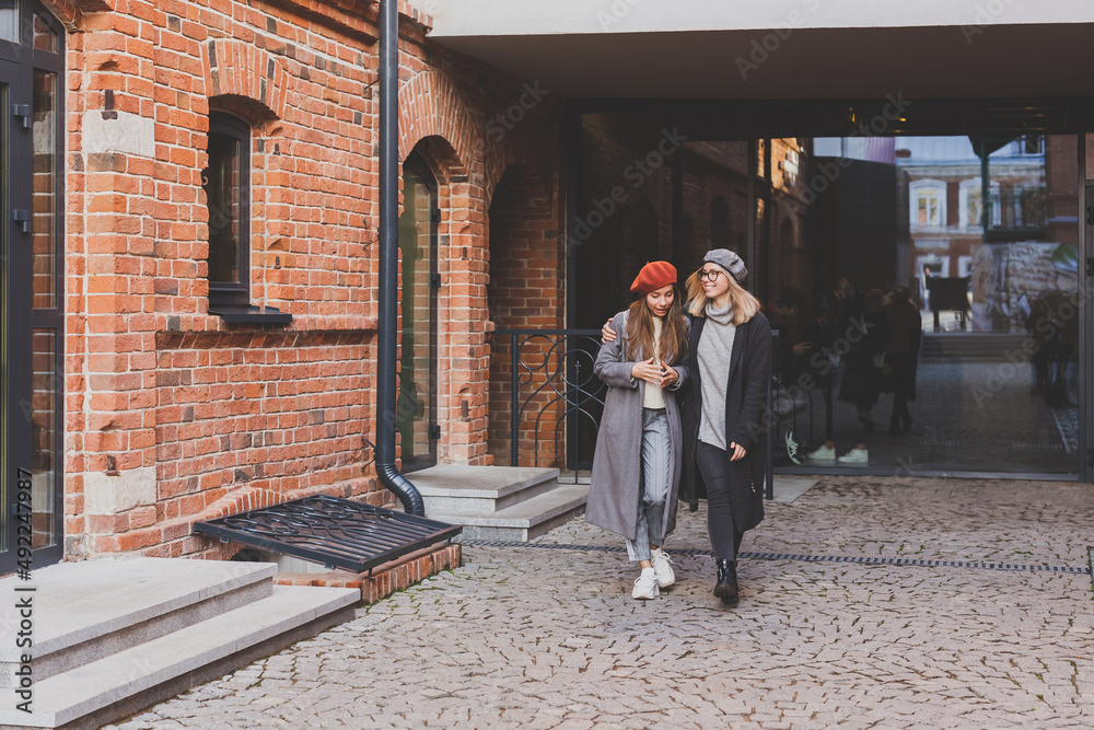 Two girls walking on street together. They are wearing spring or autumn clothes and they are happy. Friendship and relationship concept