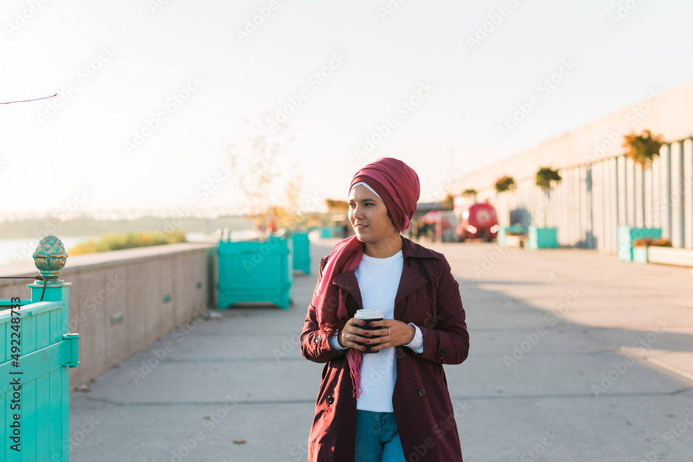 Young Arabic muslim woman in hijab walking at street seafront and drinking coffee take away - copy space and place for advertising