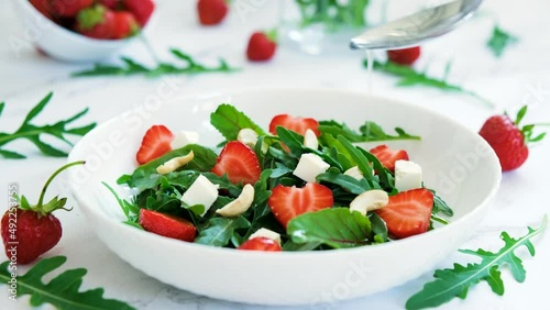 Tasty salad of strawberry with arugula, young beet tops, feta cheese and cashew nuts in white plate with salad leaves and fresh strawberry fruits around. Pouring salad with oil from spoon.