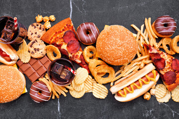 Junk food table scene scattered over a dark background. Mixture of take out and fast foods. Pizza, hamburgers, french fries, chips, hot dogs, sweets. Above view.