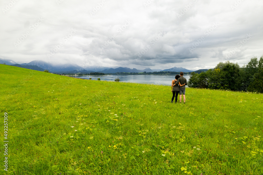 Paar umarmt sich auf einer Wiese mit See und Gebirgen im Hintergrund 