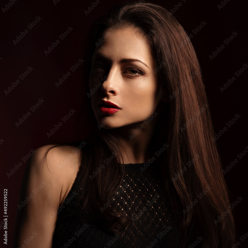 Fototapeta premium Beautiful bright makeup woman with red lipstick and long brown healthy hair with half face in shadow on dark red shadow background. Closeup