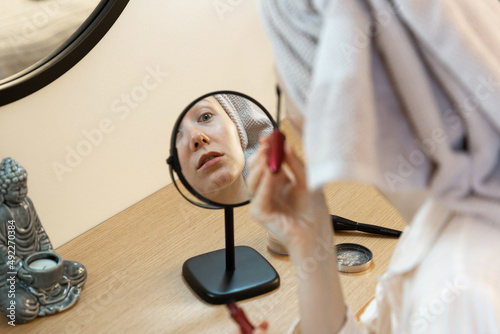 reflection in small round mirror of a young caucasian woman with wrapped hair in towel, who is applying mascara looking at mirror on makeup table, little buddha candle, beauty procedures at home