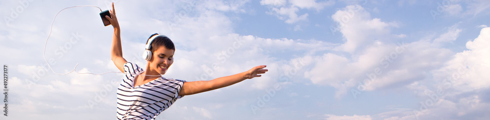 Young woman jumping on the meadow wearing headphones listening music from the smart phone on a sunny summer day