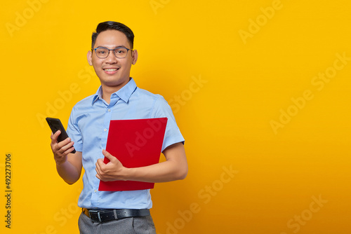 Portrait of handsome young Asian businessman in glasses standing confidently holding document folder and using mobile phone with smiling face isolated over yellow background