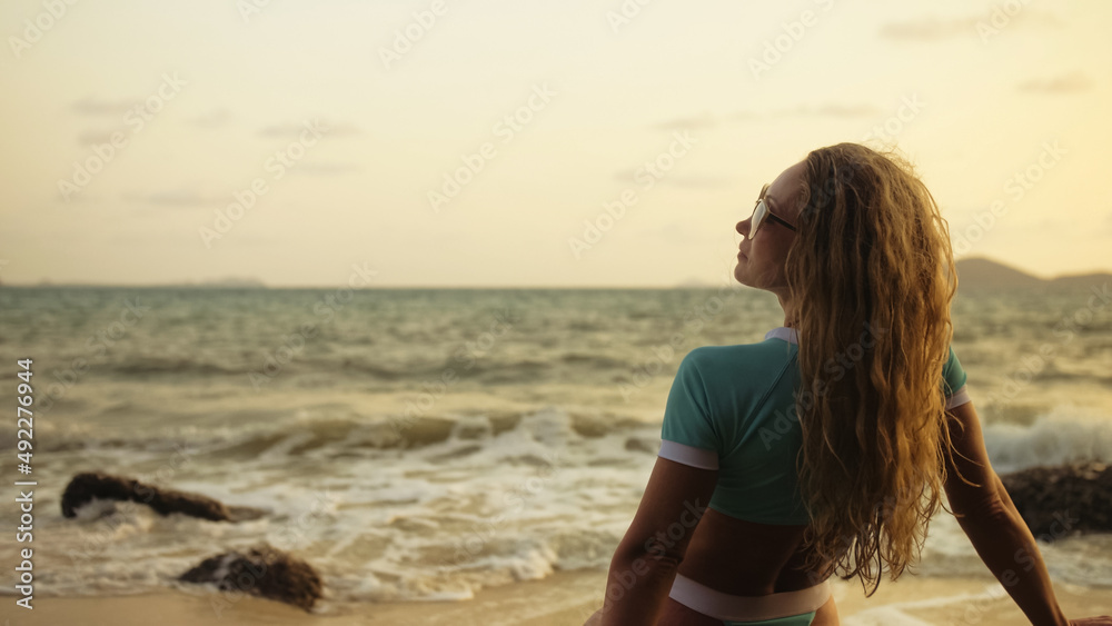 Silhouette sexy woman leaning on a rock reef enjoying and relax warm dark golden sunset. Back view. Woman in blue swimsuit and sunglasses. Concept rest tropical tourism happy summer holidays Silhouette sexy woman leaning on a rock reef enjoying and relax warm dark golden sunset. Back view. Woman in blue swimsuit and sunglasses. Concept rest tropical tourism happy summer holidays