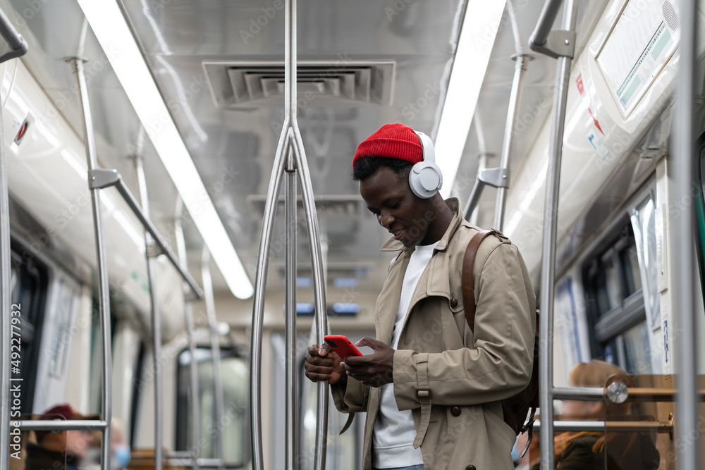 Young african american hipster man wear headphones, listening music, using smartphone in subway train. Black student male using mobile phone, chatting in social media in public transportation.