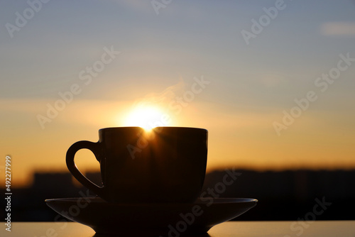 Silhouette of coffee or tea cup with steam smoke on background of sunset sky and shining sun. View from the window to evening city