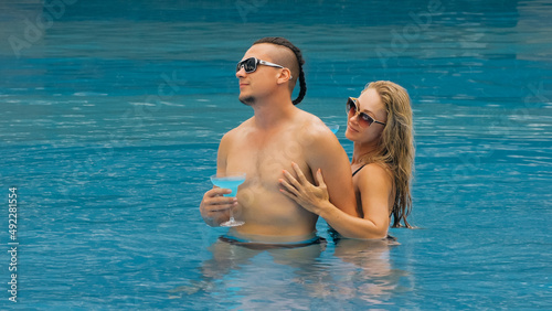 The loving couple hugs and kisses, drinking blue cocktail alcohol liquor in swimming pool at hotel outdoor. Portrait of caucasian man and woman. Creative hairstyles bodybuilder, swimsuit, sunglasses. © ivandanru
