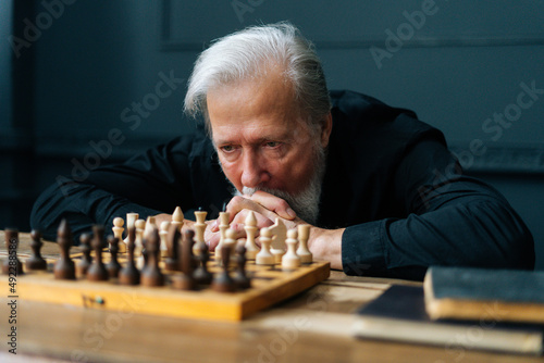 Close-up of pensive gray-haired senior older man thinking game strategy sitting on wooden table with chess board. Thoughtful bearded aged male playing chess alone at home.
