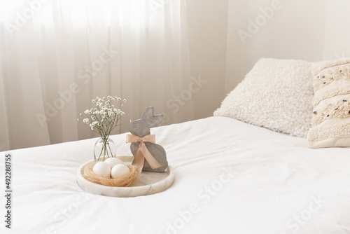 Marble tray, nest with white eggs, decorative bunny figure and vase with gypsophila flowers. White bedroom with Easter decoration. 