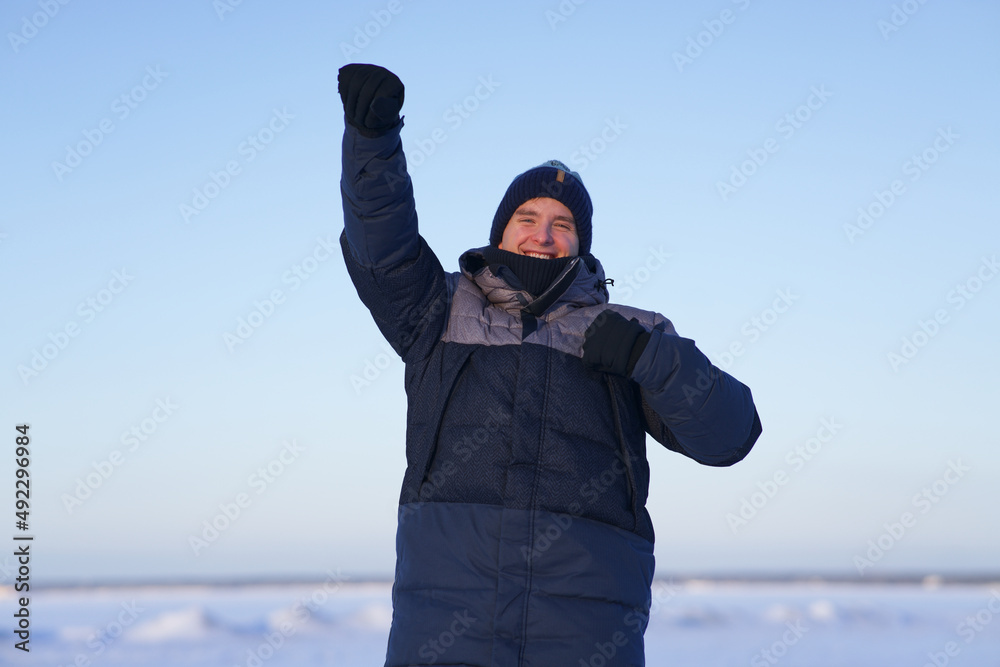 Portrait of happy successful positive guy, young cheerful man is celebrating success, victory raising up his hand and smiling at winter cold frosty snowy sunny day outdoors in snow, sky background 