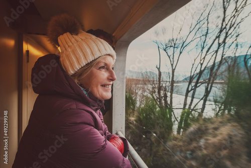 Woman (caucasian, 50s) wearing a puffer coat and warm hat, looking out a train viewing carriage 