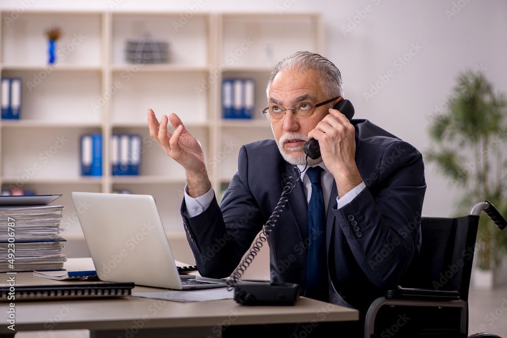 Old male employee in wheel-chair sitting at workplace Old male employee in wheel-chair sitting at workplace